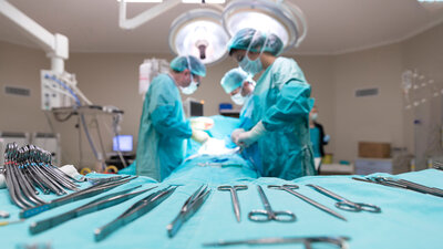 OP-Besteck liegt während einer Operation bereit, im Hintergrund operieren mehrere Menschen unter einer Leuchte.