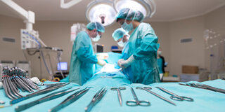 OP-Besteck liegt während einer Operation bereit, im Hintergrund operieren mehrere Menschen unter einer Leuchte.