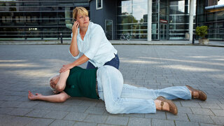 Eine Frau liegt in der Seitenlage auf dem Boden, eine andere kniet mit Handy am Ohr neben ihr