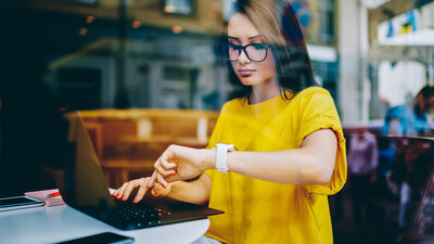 Junge Frau mit Brille schaut auf ihre Uhr während sie am Laptop sitzt