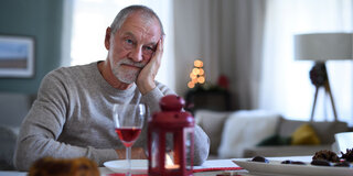 Ein älterer Mann stützt seinen Kopf auf die linke Hand und blickt nachdenklich über ein halbleeres Weinglas in die Ferne.