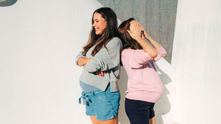 Zwei schwangere Frauen stehen Rücken an Rücken, eine hält sich die Augen zu, die andere lacht.