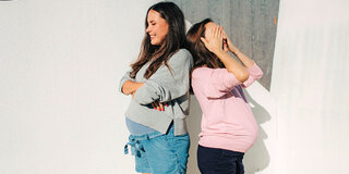 Zwei schwangere Frauen stehen Rücken an Rücken, eine hält sich die Augen zu, die andere lacht.