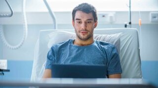 Ein Mann sitzt in einem Krankenhausbett und blickt auf ein Notebook.