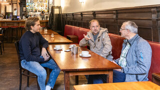 Drei Männer sitzen in einem Wirsthaus am Tisch und reden miteinander, vor ihnen stehen Kaffetassen.