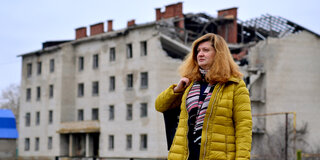 Eine Frau in einem Wintermantel blickt in die Ferne, im Hintergrund ist ein zerstörtes Mehrfamilienhaus zu sehen.