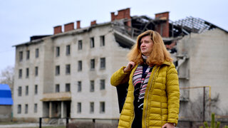 Eine Frau in einem Wintermantel blickt in die Ferne, im Hintergrund ist ein zerstörtes Mehrfamilienhaus zu sehen.