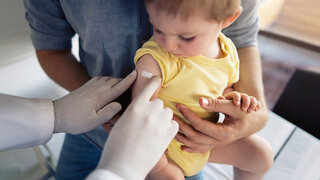 Ein Arzt klebt einem Kleinkind ein Pflaster auf den rechten Oberarm.