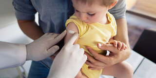 Ein Arzt klebt einem Kleinkind ein Pflaster auf den rechten Oberarm.