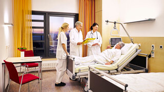 In einem Raum im Krankenhaus stehen mehrere Betten mit Patienten und medizinisches Personal läuft herum.