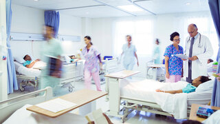 In einem Raum im Krankenhaus stehen mehrere Betten mit Patienten und medizinisches Personal läuft herum.