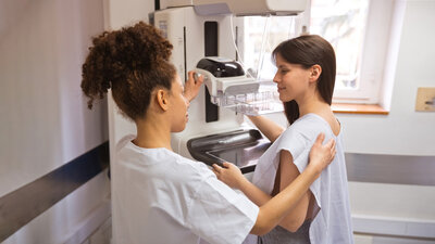 Eine medizinische Fachangestellte erklärt einer Patientin vor einem Mammographiegerät, wie die Untersuchung abläuft.