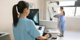 Eine medizinische Fachangestellte steuert aus einiger Entfernung ein Mammographiegerät, mit dem gerade eine Patientin untersucht wird.