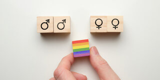 Eine Hand legt ein Bauklötzchen mit einer Regenbogenfahne zwischen weitere Bauklötzchen mit Symbolen für gleichgeschlechtliche Partnerschaften.
