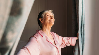 Eine ältere Frau öffent das Schlafzimmerfenster und blickt lächelnd nach draußen.