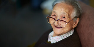 Eine ältere Dame blickt über den Rand ihrer Brille in die Kamera und lächelt.