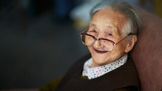 Eine ältere Dame blickt über den Rand ihrer Brille in die Kamera und lächelt.