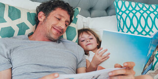 Ein Vater liegt neben seinem Kind im Bett und liest aus einem Buch vor.