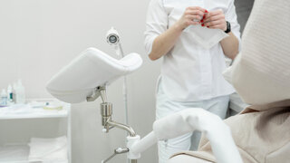 Eine Frau sitzt auf einem gynäkologischen Stuhl und eine Ärztin in weißem Gewand steht vor ihr.