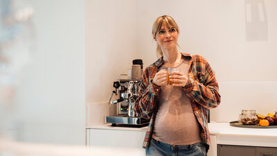 Eine Schwangere lehnt an einem Küchentresen und hält einen Becher in der Hand.