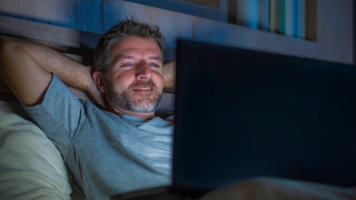 Ein Mann sitzt in einem dunklen Raum und schaut auf einen hell leuchtenden Notebook-Bildschirm.