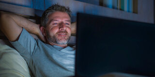 Ein Mann sitzt in einem dunklen Raum und schaut auf einen hell leuchtenden Notebook-Bildschirm.