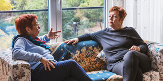 Zwei Frauen sitzen auf einem Sofa und diskutieren miteinander, eine der beiden gestikuliert, die andere schaut skeptisch.