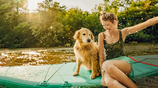 Eine Frau sitzt mit einem Hund auf einem Surfbrett und spritzt mit der Hand Wasser in die Höhe.
