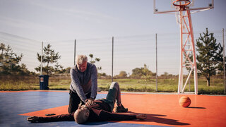 Ein Mann reanimiert einen anderen Mann auf einem Basketballplatz mit einer Herzdruckmassage.
