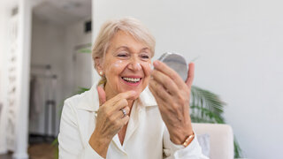 Eine Frau schaut in einen Handspiegel und trägt Creme im Gesicht auf.
