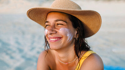 Eine lächelnde Frau mit Sonnenhut und Sonnencreme auf den Wangen sitzt am Strand.