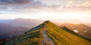 Auf einer Bergkuppe führt ein schmaler Weg Richtung Horizont, in der Ferne geht die Sonne auf.