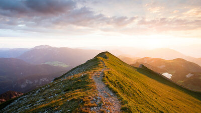 Auf einer Bergkuppe führt ein schmaler Weg Richtung Horizont, in der Ferne geht die Sonne auf.