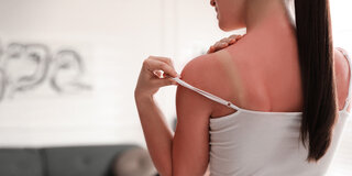 Eine Frau mit einem Sonnenbrand hebt den Träger ihres Tops an, sodass sich darunter ein weißer Streifen auf der Haut zeigt.