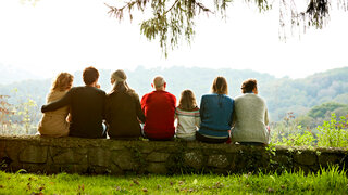 Jüngere und ältere Menschen sitzen nebeneinander auf einem Baumstamm und blicken in die Ferne.