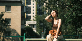 Eine Frau sitzt im Sonnenschein auf einer Bank und wischt sich den Schweiß mit einem Tuch von der Stirn.