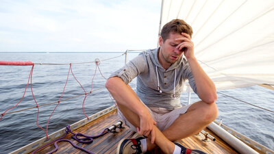 Ein erschöpfter Mann sitzt auf einem Segelboot und hält sich eine Hand an den Kopf. 