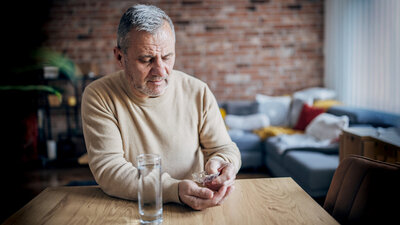 Älterer Mann sitzt an einem Tisch, will eine Tablette nehmen. Daneben steht ein Glas Wasser. 