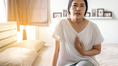 Junge Frau sitzt auf dem Bett und drückt Hand gegen das Brustbein