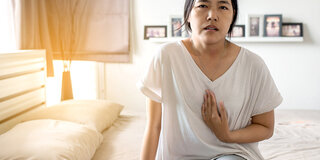 Junge Frau sitzt auf dem Bett und drückt Hand gegen das Brustbein