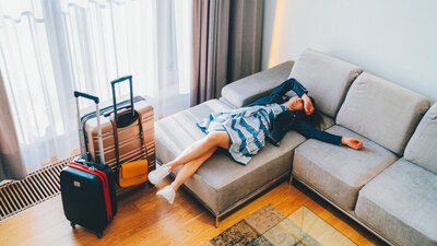 Eine Frau liegt erschöpft auf dem Sofa eines Hotelzimmers, neben sich zwei Koffer.