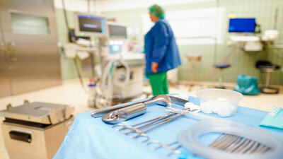 In einem Operationssaal liegen chirurgische Instrumente und im Hintergrund steht eine Frau im Kittel vor Monitoren