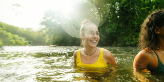 Zwei Frauen beim Baden in einem Naturgewässer. Im Hintergrund sind Bäume zu sehen.