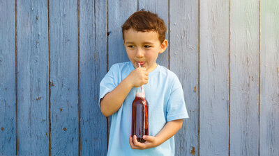 Ein Junge in blauem T-Shirt steht vor einer Wand und trinkt mit einem Strohalm aus einer Flasche.